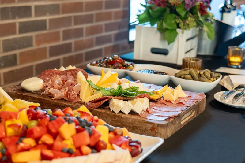 A charcuterie board of prosciutto, salami, brie and cheddar beside a fruit platter, olives and cornichons on a black-draped buffet