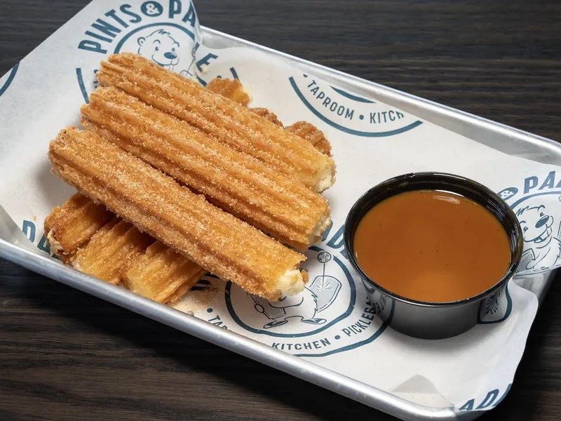 Four cinnamon-sugar churros on a branded tray with a ramekin of warm caramel dipping sauce