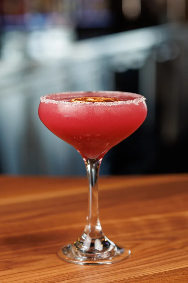A deep pink cocktail in a sugar-rimmed coupe glass with a dried citrus wheel floating on top, on the wood bar
