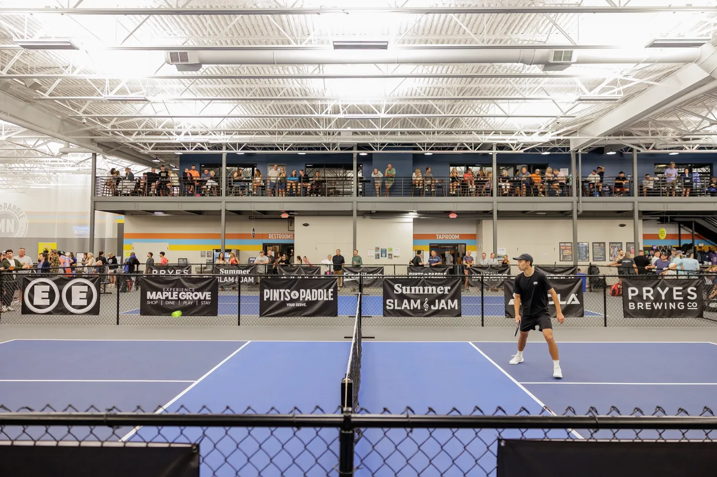 A wide view of the indoor courts during a tournament: a player at the net, banners along the fences and a full crowd on the mezzanine