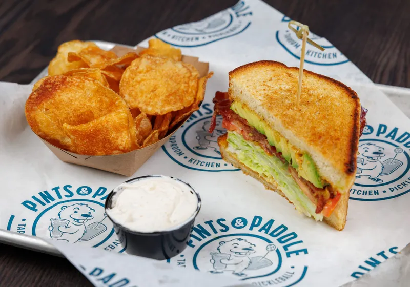 Half an avocado BLT on toasted bread with a boat of kettle chips and a cup of dip on a Pints & Paddle tray