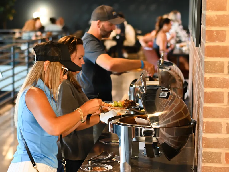 Two guests in ball caps filling plates at a row of stainless chafers on the mezzanine, more guests at the tables behind them