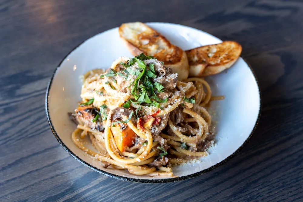 A bowl of short rib bucatini pasta topped with grated Parmesan and basil, with two garlic crostinis.